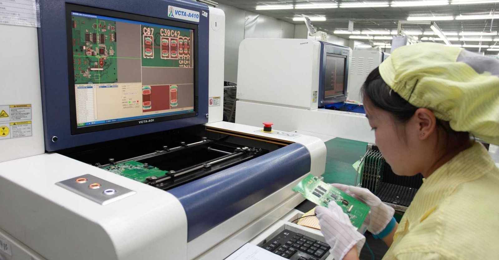 AOI review workstation example | GlobalWin Operator inspecting a circuit board beside an optical-inspection monitor and workstation